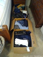 Two cardboard boxes are shown side by side on a carpeted floor next to a wooden bed and dresser. The boxes contain folded men's clothing in various colors and patterns, including shirts, sweatshirts, and pajamas. Some winter gloves and scarves are visible.