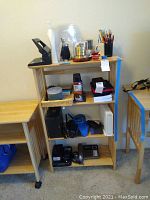 Wooden shelving unit with office supplies, electronic devices, and phone equipment arranged across three shelves. Items include routers, a landline phone, Blackberry phone, blank CDs, pens, pencils, three-hole punch, cases for tablet and Samsung Galaxy S8, and other miscellaneous office supplies.