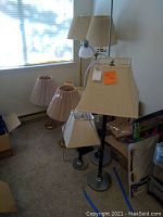 Photo showing five assorted lamps including two tall brass base floor lamps with pleated beige shades, a tall silver base floor lamp with rectangular beige shade, and two desk lamps with silver bases and small white and beige shades.