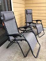 Two gray antigravity folding lounge chairs shown unfolded, positioned outside on a concrete floor by a building with yellow siding.