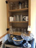 Photo showing wooden cabinet interior with coffee mugs, clear glass mugs, antique hurricane lamp on counter, and decorative glass bowls in front.