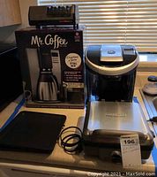 Photo showing Mr. Coffee 10-cup programmable coffee maker in original boxed packaging, Keurig coffee maker, and Cuisinart Belgian waffle maker on countertop with power cords visible.