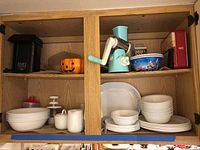 Two wooden cabinet shelves containing Corelle bowls, plates, cups, ceramic containers with lids, rotary grater, salad shooter, coffee grinder, and popcorn popper.