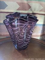 Front angle of the large purple and white swirled handkerchief vase on a metal table with a patterned fabric background.