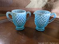 Side view of blue hobnail opalescent glass creamer and sugar bowl set on a dark brown table.