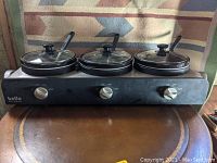 Front view of the Bella Kitchen triple slow cooker with three ceramic pots and glass lids, each with a spoon inside, sitting on a brown surface with a patterned cloth backdrop.