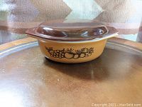 Side view of oval casserole dish showing Old Orchard fruit pattern with grapes and apples on golden-orange base, brown lid on top.