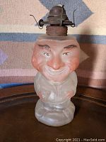Front view of the figural oil lamp showing the red-faced man with smiling expression and metal burner on top.