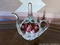 Front angle view of clear glass teapot-shaped paperweight with red flowers inside on brown surface with patterned background.