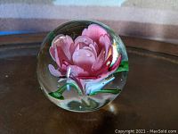 Clear spherical glass paperweight with a single pink flower blossom inside. The flower shows detailed petals and green leaves at the base. The glass is smooth and clear.