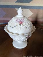 Full view of Westmoreland glass candy dish with lid on a pedestal showing painted pink roses, purple bows, and blue flowers on white milk glass embossed with grape and fruit motif.