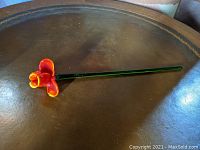Side view of a long green stem supporting an open glass flower with orange and yellow petals.
