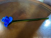 Full view of the long stemmed blue glass flower lying on a brown surface showing the entire length and shape.