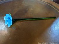 Full view of the blue glass flower with a long green stem on a brown surface.