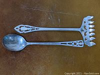 Top view of the sterling silver sugar spoon and sardine fork lying on a brown surface, showing pierced decorative handles and overall shape of each piece.