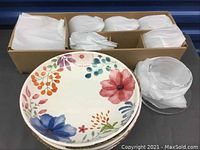 Floral microwaveable dinner plates stacked in front of boxed glassware plates and cups wrapped in paper.