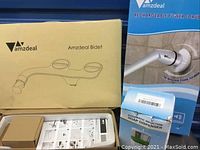 Photo showing Amzdeal bidet packaging and internal components in open box, with rechargeable power scrubber and automatic soap dispenser packaging in background.