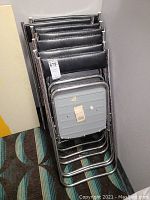 Stack of seven black padded folding chairs folded and nested against a wall.