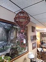 Full view of hanging wire and wooden hot air balloon with visible artificial purple flowers inside wooden cage bottom.