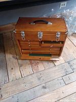 Closed wooden machinist's tool chest showing metal reinforced corners, three metal latches, and black handle on top.
