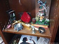 Shelf display showing multiple items including red small purse, crucifix, books, bracelet, metal bell, Trick Dog coin banks with figurines, and other small decorative objects