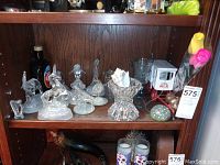 Shelf with various glass animal figurines, clear glass paperweight and a miniature milk cart model accompanied by artificial flowers.