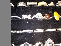 Seven sterling silver rings displayed on black velvet, showing red and yellow stones on the bottom row including a dragonfly design and pearl-like bead