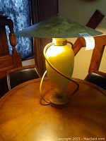 Full view of vintage table lamp on wood table, showing the tall beige ceramic base with metal coil and wide, conical, textured green metal lampshade.