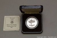 The silver two-dollar commemorative coin inside its original black display box with cream lining, showing the Bermuda cedar tree design and the certificate of authenticity card.