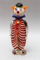 Front view of red and white striped blown glass clown figurine with orange hair, black hat, and purple collar