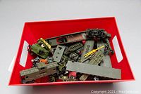 Overhead view of multiple green die cast army vehicles including tanks, U.S. Army train cars, a gray metal train car roof, and yellow plastic crane arm parts in a red bin.