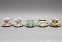 Five pairs of assorted fine porcelain teacups and saucer sets displayed side by side on neutral background, showing various floral and gold rimmed designs.