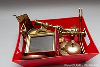 Wide view of the lot in red container showing multiple brass items including candle holders, bell, horseshoe, magnifying glass, and others.