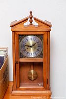 Full front view of wooden German wall clock with closed glass door, clock face, and wooden top finial.