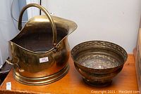 Photo showing large brass coal scuttle and decorative bowl side by side on wooden surface.