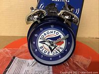 Front close-up of the Toronto Blue Jays metal alarm clock showing the team logo on the clock face with twin metal bells on top.