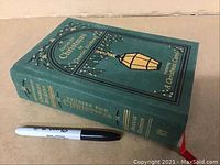 Green cloth hardcover book titled "Stories for Christmas" by Charles Dickens, showing front and spine with gold embossed text and decoration, placed next to a marker for size reference.