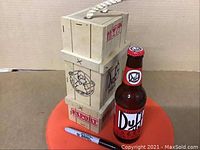 Full view of Duff Beer bottle standing next to the stackable wooden crate with a rope handle, and a Sharpie pen for scale.