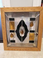Frontal image of a square stained glass window panel framed in light wood, showing the multi-colored glass design featuring black, amber-yellow and clear textured glass with hanging amber drops.