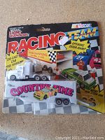 Front view of Racing Champions diecast hauler truck toy in sealed packaging showing white hauler truck with opening door ramp and yellow mini stock car included.