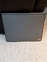 Grey portfolio case shown closed on a carpeted floor, with a visible handle and a label on the bottom corner.