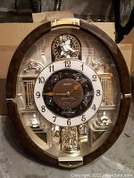 Front view of oval Seiko musical wall clock showing dial, frame, architectural backdrop and crystal pendulum