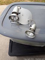 Front view of silver-tone metal 2-light LED fixture with adjustable lamp heads and wiring visible, placed on plastic bin lid.