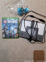 Xbox One Lego Dimensions game case with disc, two platform bases with attached cords, and bag of clear blue Lego game pieces on carpet.