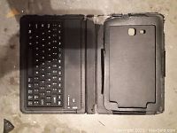 Opened view of black leather-like folio case with Bluetooth keyboard on left side and molded plastic holder for Samsung tablet on right side, showing keyboard layout and slots for tablet buttons and camera.