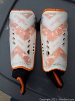 Pair of white and orange kids soccer shin pads with ventilation holes and elastic straps.
