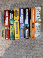 Photo showing nine hardcover Clive Cussler books arranged on a carpeted surface. Titles on spines are readable and indicate variety in his adventure novels.