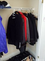 Six coats hanging on a closet rod including blue, black, red, gray and dark coats above footwear