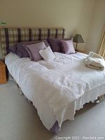 Full view of king size bed with plaid upholstered headboard, white quilting coverlet, purple pillows, and beige throw on top