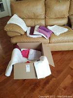 Box and sofa with assorted folded white, purple, and pink linens including tablecloths, placemats, and napkins.